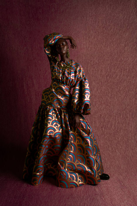 Elegant black woman poses wearing a patterned headscarf and voluminous matching gown in a studio with a textured mauve backdrop.