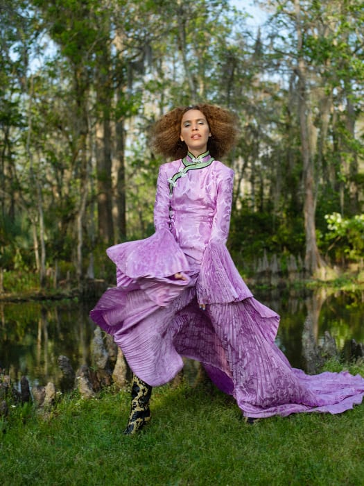 A woman in an elaborate purple dress poses in a lush, green outdoor setting near a body of water with trees reflecting in the water.