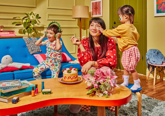 A mother sits at a table with her children, one painting her face, with a cake, flowers, couch, and various objects visible.