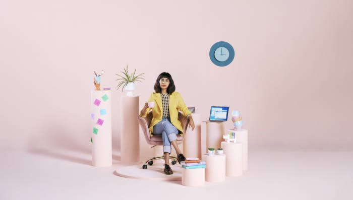 Woman in yellow blazer sitting in a chair holding a mug, surrounded by desks, potted plant, and computer against a pink background.