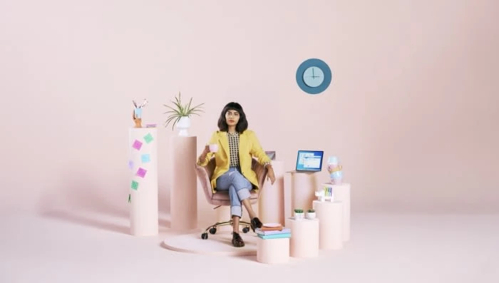 Woman in yellow blazer sitting in a chair holding a mug, surrounded by desks, potted plant, and computer against a pink background.