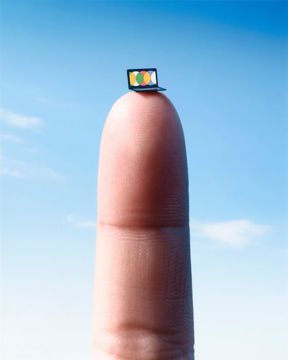 A miniature laptop sits atop a person's finger against a vibrant blue sky, creating a whimsical and tech-focused visual composition.