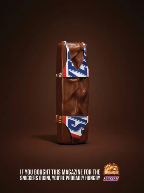 Snickers candy bar is depicted wearing a bikini with Snickers logo against a brown background in a promotional advertisement.