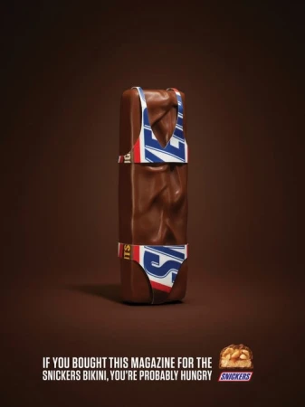 Snickers candy bar is depicted wearing a bikini with Snickers logo against a brown background in a promotional advertisement.