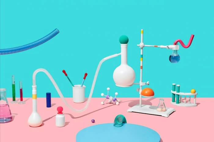 Still life of laboratory glassware and equipment on a pink surface against a turquoise background featuring test tubes and beakers.