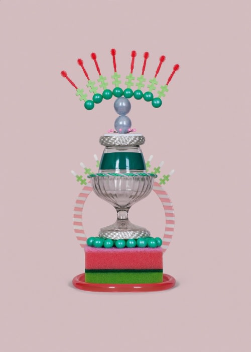 Still life photography featuring a whimsical tiered sculpture made of everyday objects like glass bowls, sponges, and beads on a light pink backdrop.