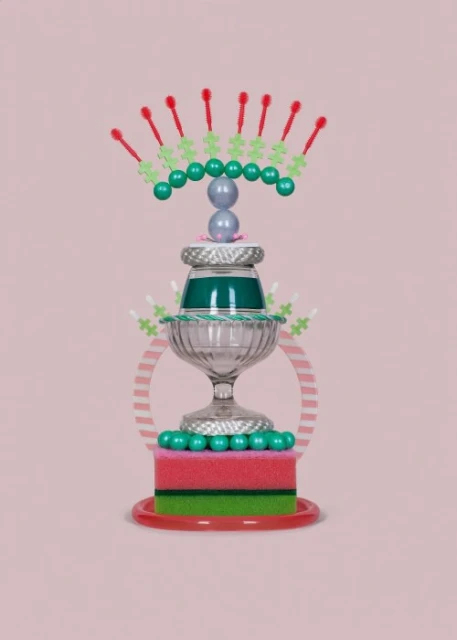 Still life photography featuring a whimsical tiered sculpture made of everyday objects like glass bowls, sponges, and beads on a light pink backdrop.