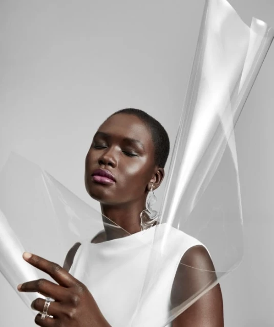 Portrait of a woman with dark skin and closed eyes wearing a white top with earrings, styled with a translucent plastic sheet.