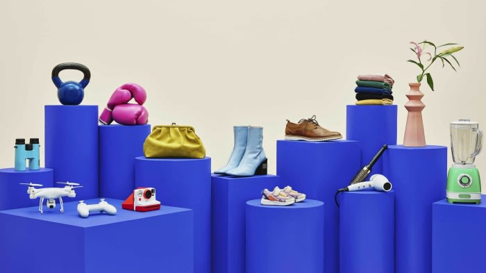 Arrangement of diverse objects including sports equipment, clothing, shoes, consumer electronics, and home goods on blue pedestals.
