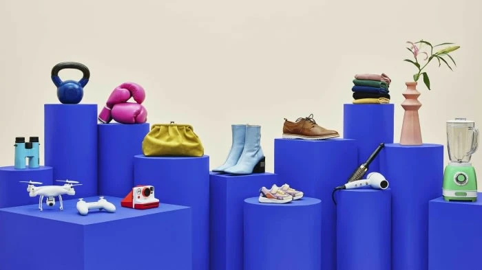 Arrangement of diverse objects including sports equipment, clothing, shoes, consumer electronics, and home goods on blue pedestals.