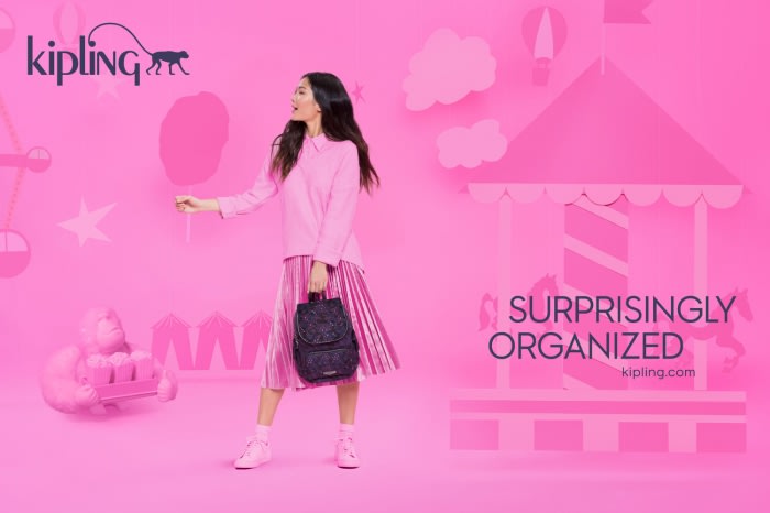 Asian woman in pink outfit holding backpack, standing near cut-out carousel. Pink backdrop with Kipling logo. Beauty shot, studio lighting.