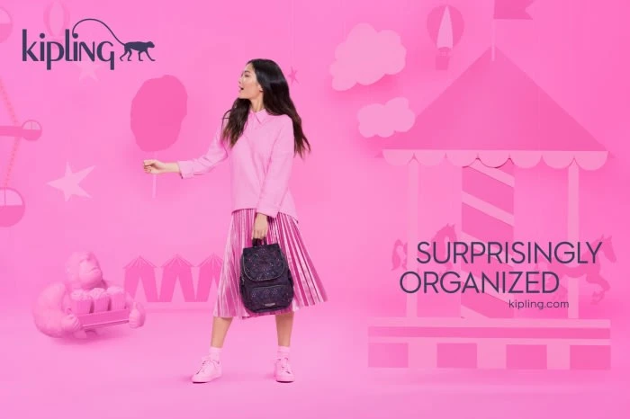 Asian woman in pink outfit holding backpack, standing near cut-out carousel. Pink backdrop with Kipling logo. Beauty shot, studio lighting.