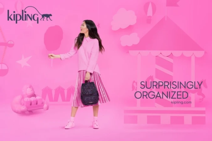 Asian woman in pink outfit holding backpack, standing near cut-out carousel. Pink backdrop with Kipling logo. Beauty shot, studio lighting.