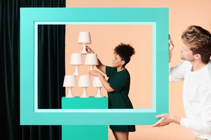 Two people pose with a tower of table lamps inside a turquoise frame against a split background, indoor, product shot.