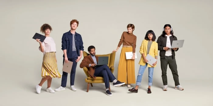 Group of six young adults using laptops in a neutral studio, dressed in casual wear. One person is sitting in an armchair.