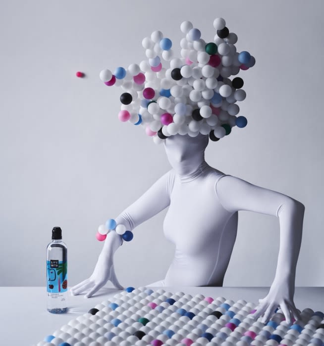 Person in white bodysuit sits at a table with bottle and beaded objects, white background, creatively styled in surreal composition.