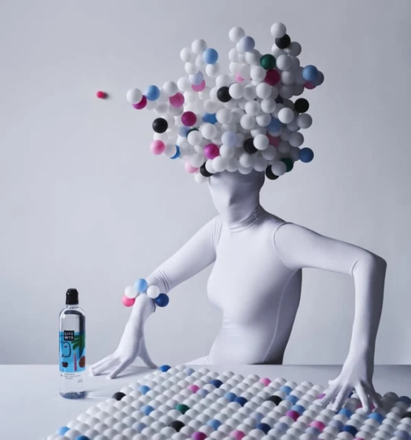 Person in white bodysuit sits at a table with bottle and beaded objects, white background, creatively styled in surreal composition.