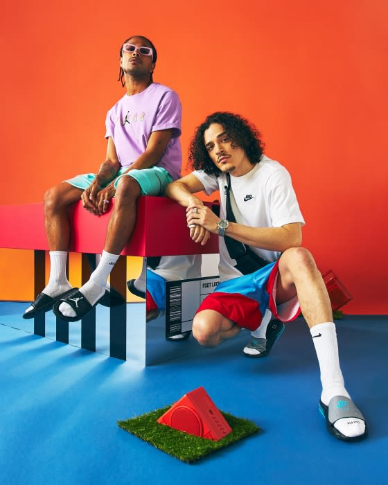 Two adult males pose in a studio setting, one seated on a block and the other squatting, wearing casual athletic apparel and slides.