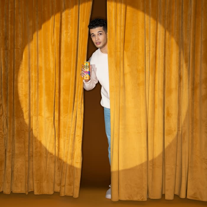 A young adult stands behind a gold curtain, holding a beverage can in a brightly lit studio with a neutral brown background.
