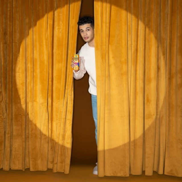 A young adult stands behind a gold curtain, holding a beverage can in a brightly lit studio with a neutral brown background.
