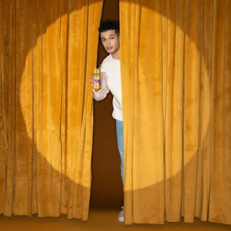 A young adult stands behind a gold curtain, holding a beverage can in a brightly lit studio with a neutral brown background.