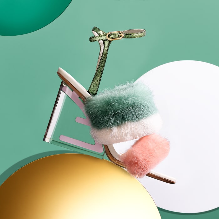 Elevated studio shot of a single green, white, and pink high-heeled sandal with a clear heel on a multicolored circular background.