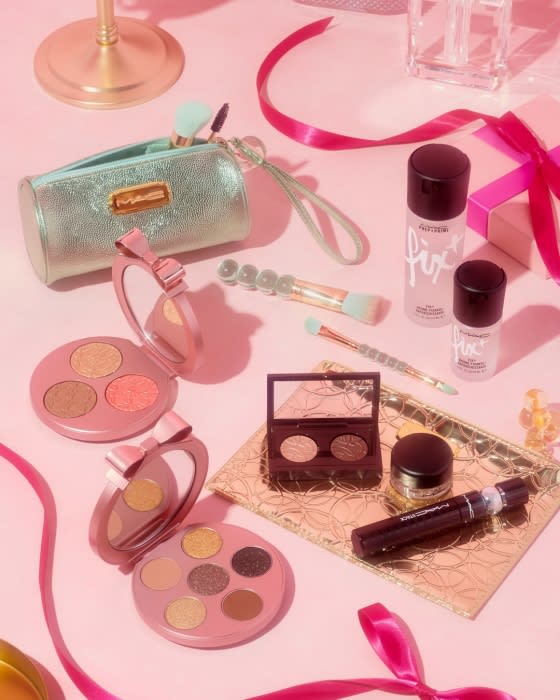 Cosmetic product arrangement on pink background: eyeshadow palettes, makeup brushes, setting spray, and pink ribbons.
