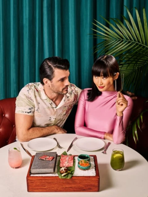 A stylish couple sits at a restaurant table with pink and green drinks, ready to enjoy gourmet meat on a wooden platter.