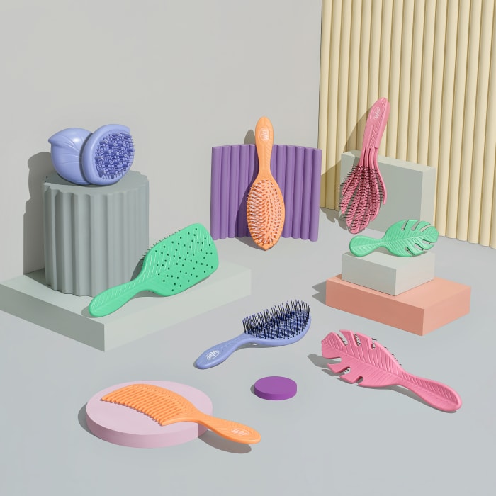 Assorted colourful hairbrushes and combs are displayed on a light gray background with pastel platforms and accents in a product shot.