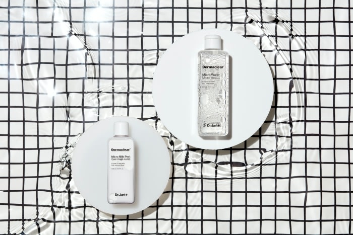 Two bottles of DermaClear skincare product on white circular stands, photographed from above against a black-and-white checkered backdrop.