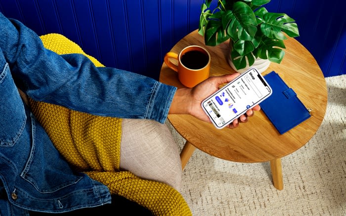 Person holds smartphone showcasing online shopping app near coffee, notebook, and plant on wooden table against blue wall backdrop.