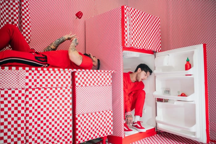 Two men in red outfits pose in a red and white checkered room with a refrigerator, one lying down and one crouching by the fridge.