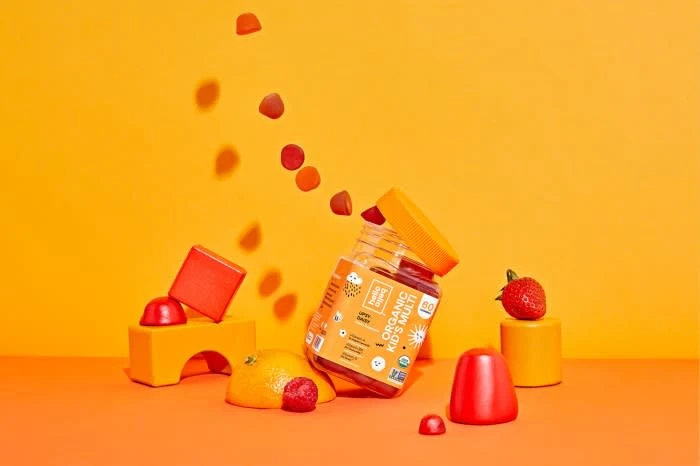 Gummy vitamins and fruits on an orange background. Vitamin bottle is surrounded by colorful candies, strawberries, and citrus fruits.