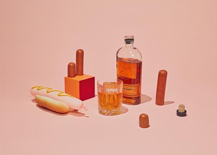 Still life shot of a hot dog with mustard, whiskey glass with ice, bourbon bottle, and sausage pieces on a pink background.