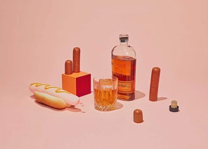 Still life shot of a hot dog with mustard, whiskey glass with ice, bourbon bottle, and sausage pieces on a pink background.