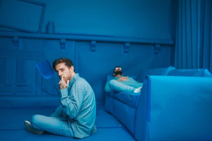 Man with tattoos sits cross-legged in a monochromatic blue room while another man relaxes on a blue couch, creating a surreal atmosphere.
