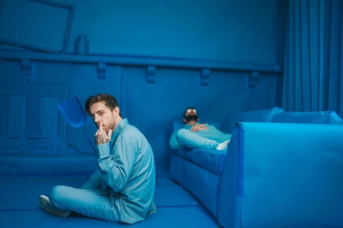 Man with tattoos sits cross-legged in a monochromatic blue room while another man relaxes on a blue couch, creating a surreal atmosphere.