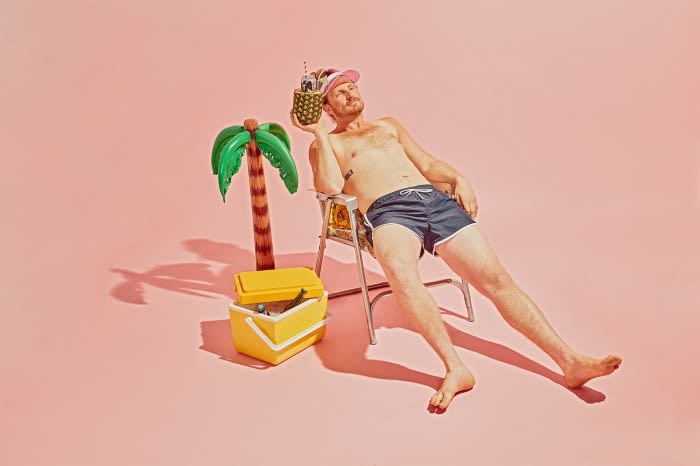 Man lounges on a beach chair holding a pineapple cocktail, with a cooler and inflatable palm tree against a pink studio backdrop.