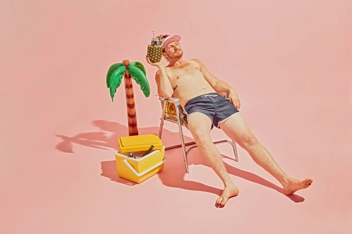 Man lounges on a beach chair holding a pineapple cocktail, with a cooler and inflatable palm tree against a pink studio backdrop.