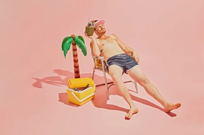 Man lounges on a beach chair holding a pineapple cocktail, with a cooler and inflatable palm tree against a pink studio backdrop.