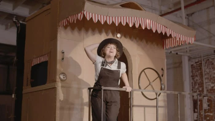 Woman stands on a stage in front of a large cardboard structure with a red and white striped awning in a warehouse-like space.