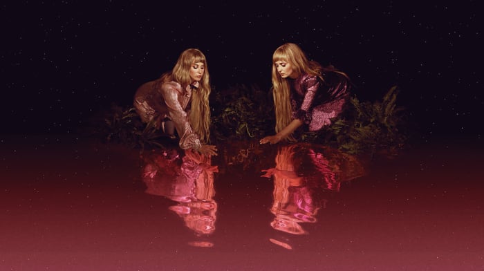 Two women with long blonde hair kneeling on a body of water. Reflections visible in red water. Dark sky with starry background.