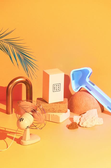 Studio shot of a box with text, a vintage fan, a copper u-bend, a blue plastic toy rake, and various geometric objects against an orange backdrop.