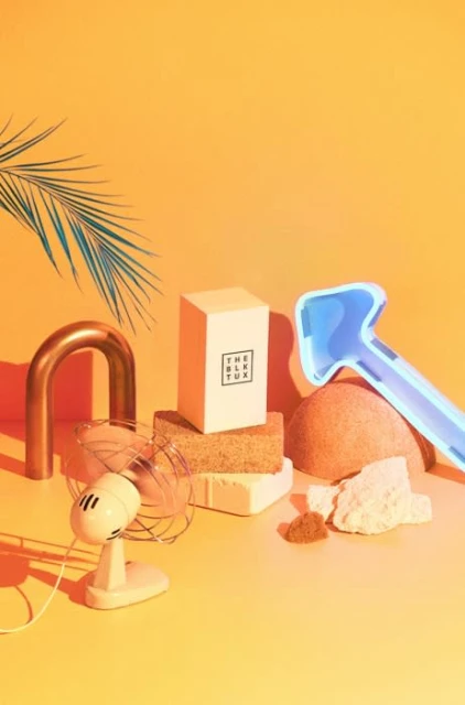Studio shot of a box with text, a vintage fan, a copper u-bend, a blue plastic toy rake, and various geometric objects against an orange backdrop.