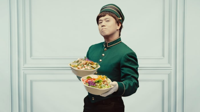 Asian man dressed as bellhop holds two salad bowls in front of a light blue paneled wall, providing a whimsical and healthy food visual.