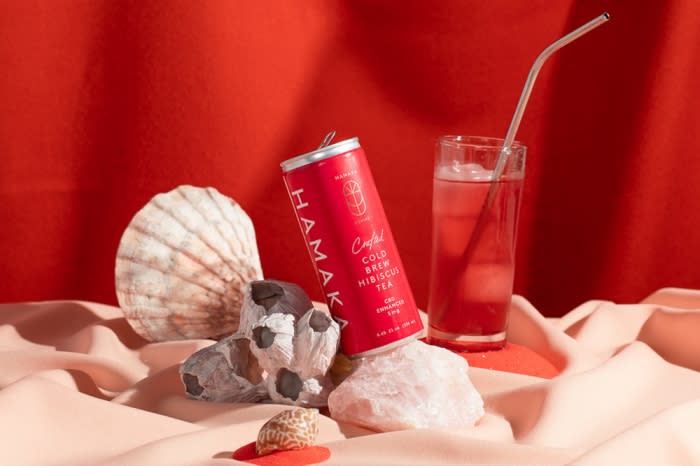Still life with a red canned drink, glass of beverage with straw, and seashells, staged on peach fabric against a red backdrop.