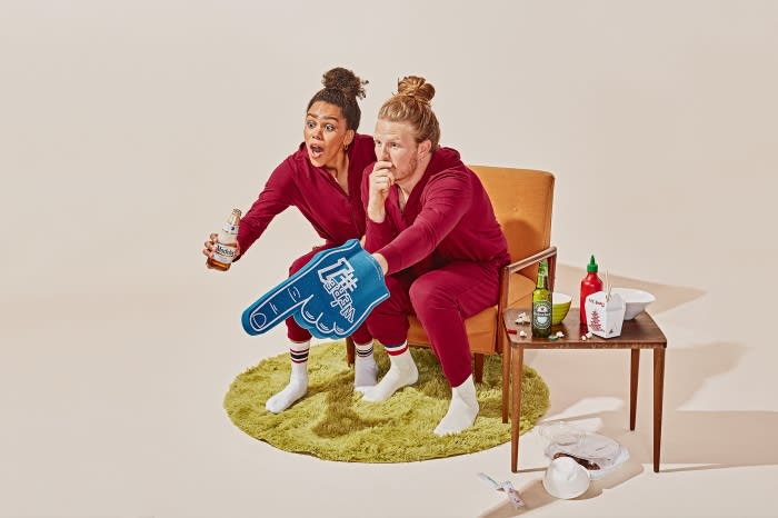 Enthusiastic couple in matching outfits cheers with a foam finger, beers, and takeout, sitting on a retro chair against a solid backdrop.