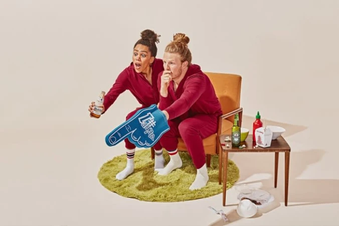 Enthusiastic couple in matching outfits cheers with a foam finger, beers, and takeout, sitting on a retro chair against a solid backdrop.