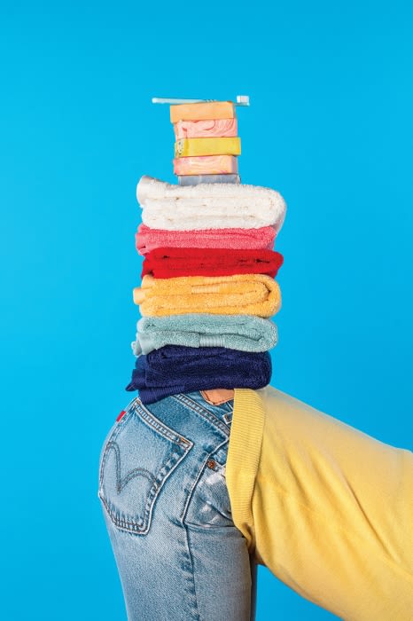 Person balancing a stack of colorful folded towels and toiletries on their head. The person is wearing denim jeans and a yellow sweater.