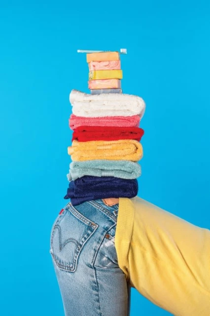 Person balancing a stack of colorful folded towels and toiletries on their head. The person is wearing denim jeans and a yellow sweater.